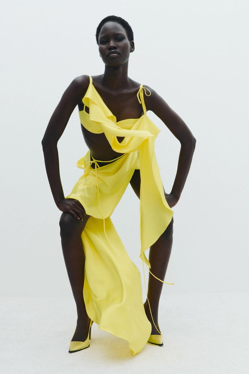 A dark-skinned woman poses confidently in a light yellow dress and matching heels against a bright white background, fashion shot.