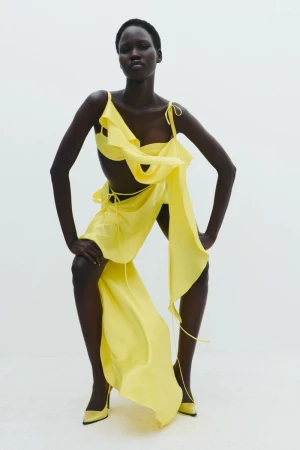 A dark-skinned woman poses confidently in a light yellow dress and matching heels against a bright white background, fashion shot.