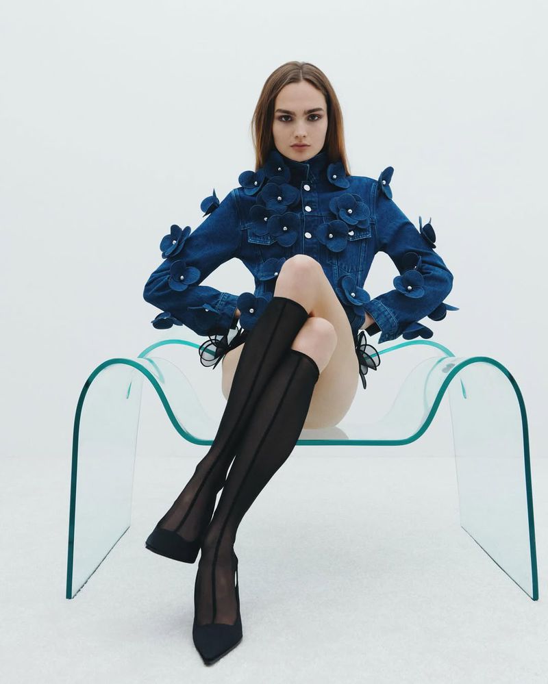 A poised female model is seated on a transparent chair in a photo studio, wearing a floral-accented denim jacket and black sheer tights with heels.