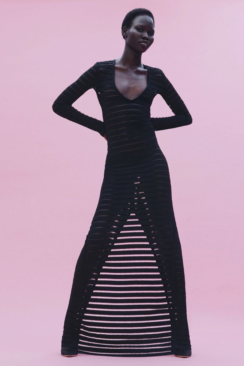 Model posing in a form-fitting black long-sleeved knit dress with front ladder cutouts against a pastel pink backdrop.
