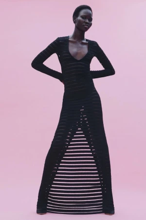 Model posing in a form-fitting black long-sleeved knit dress with front ladder cutouts against a pastel pink backdrop.