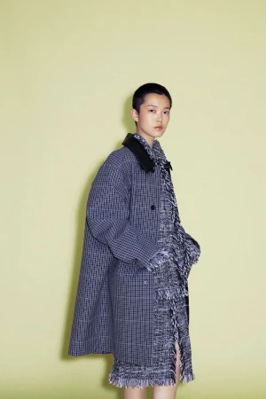 Model Echo Zhao poses in an oversized checked coat with a dark collar over a fringed tweed garment against a pale yellow studio backdrop.