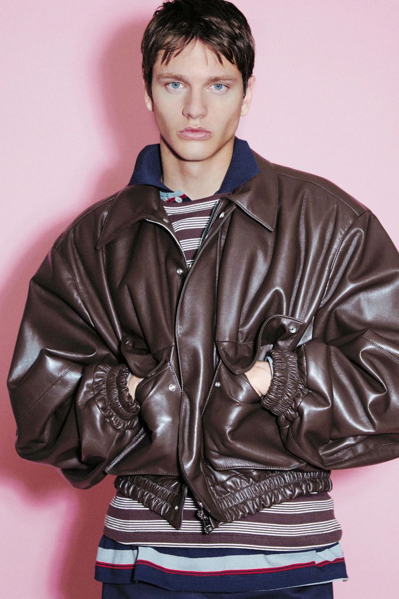 Male model posing in an oversized dark brown leather bomber jacket over a striped shirt against a pink studio background.