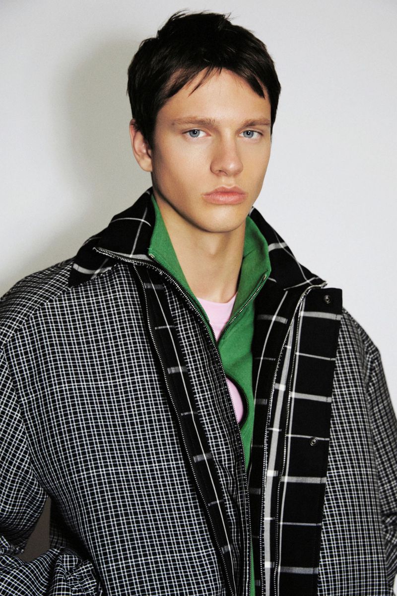 Young male model wearing a black and white grid jacket over a green zipped collar against a neutral studio backdrop.