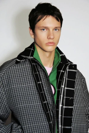 Young male model wearing a black and white grid jacket over a green zipped collar against a neutral studio backdrop.