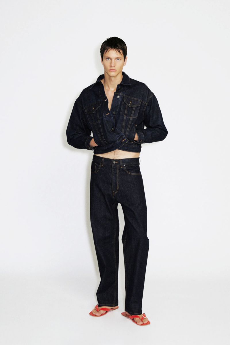 Male model wearing a dark raw denim cropped jacket and matching wide-leg jeans with red sandals against a white background.