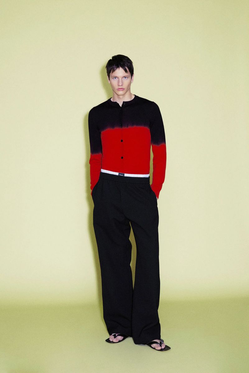 Model wearing a black and red color-blocked cardigan and black trousers against a pale yellow seamless background.