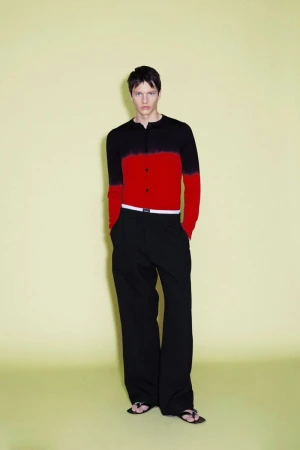 Model wearing a black and red color-blocked cardigan and black trousers against a pale yellow seamless background.