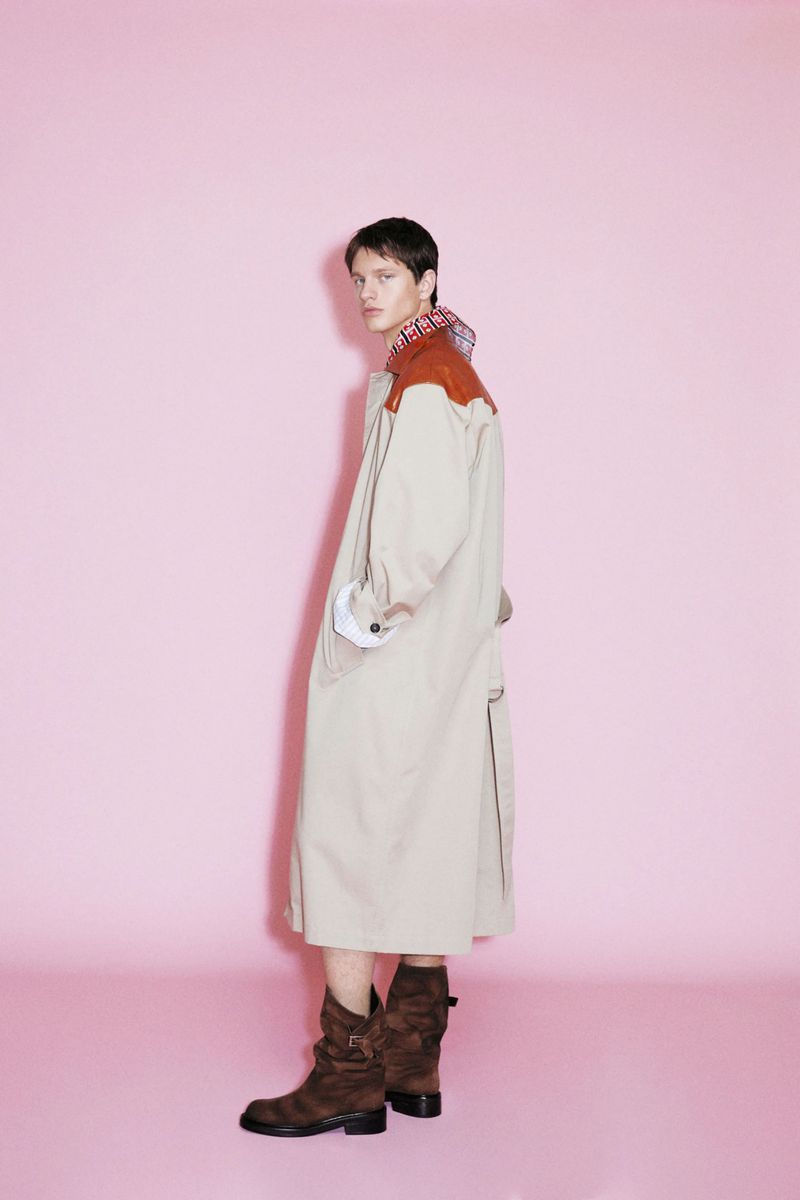 Model wearing a long beige trench coat with rust-colored shoulders and brown boots against a pink background