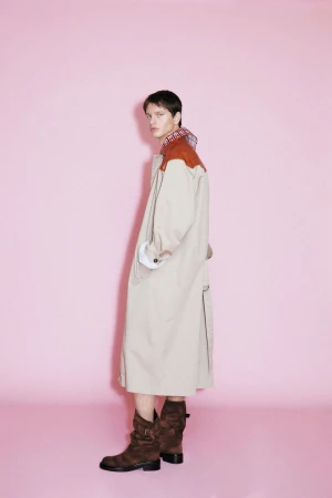 Model wearing a long beige trench coat with rust-colored shoulders and brown boots against a pink background