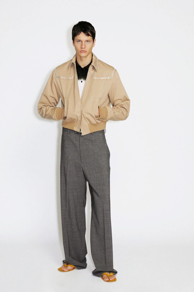 Model wearing a beige embellished zip jacket and wide-leg grey trousers against a white seamless backdrop.