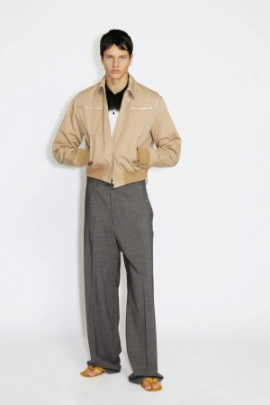 Model wearing a beige embellished zip jacket and wide-leg grey trousers against a white seamless backdrop.