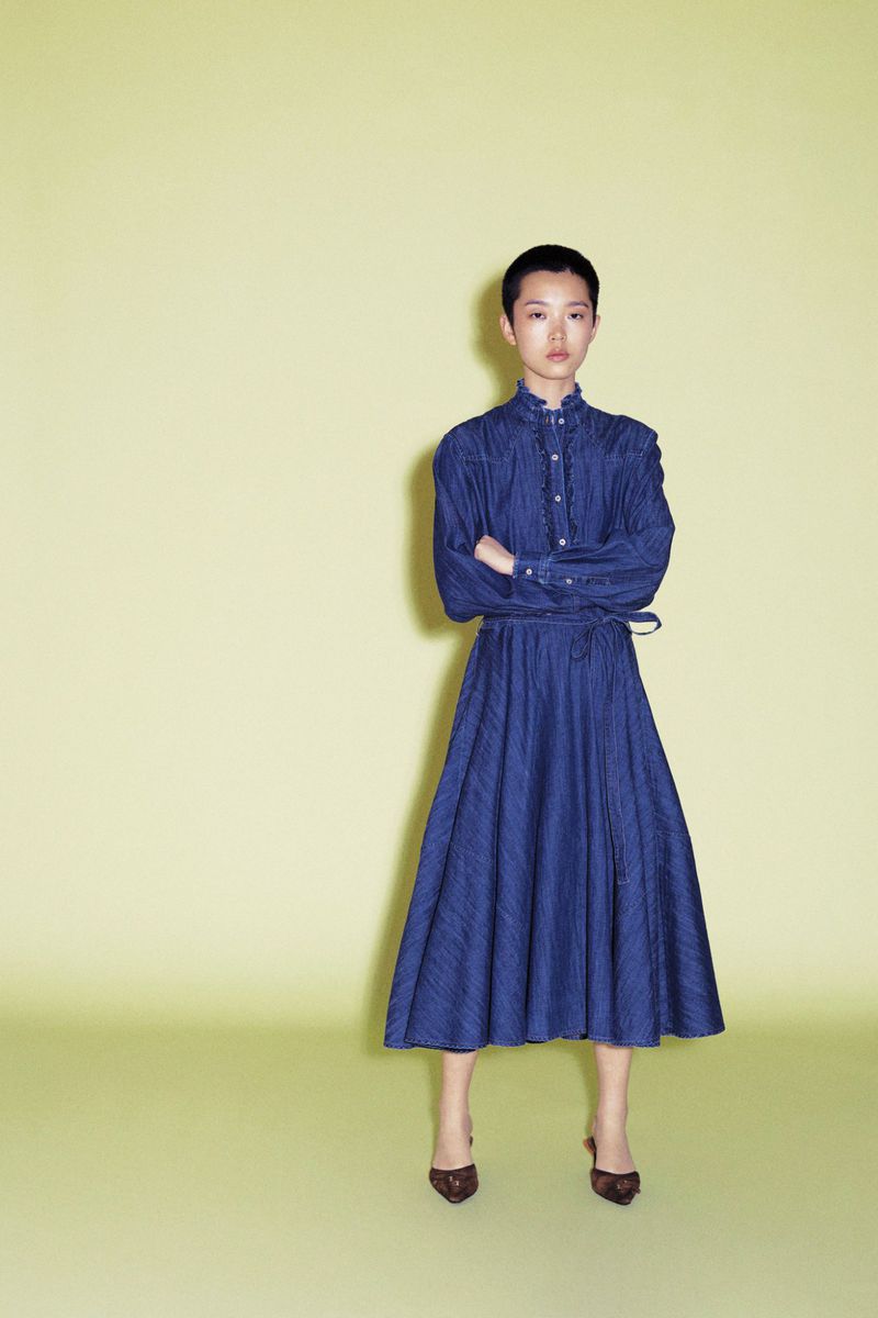 Model Echo Zhao wearing a long-sleeve blue belted midi dress with arms crossed against a pale yellow-green background.