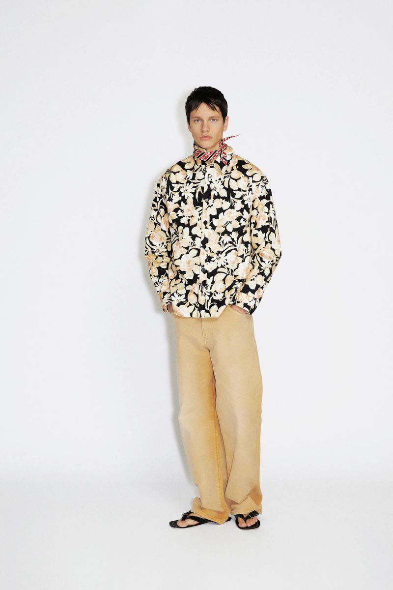 Model wearing a long-sleeve floral print shirt, patterned neck scarf, and wide-leg beige trousers against a white seamless backdrop.