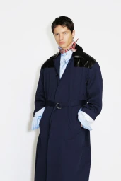 Model wearing a navy blue belted coat with black shoulder panels and a patterned neck scarf against a white studio background.