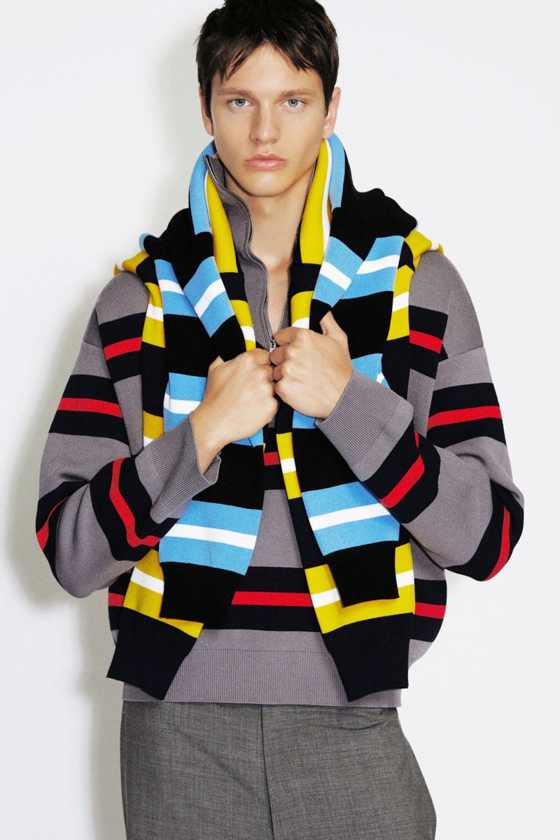 Model wearing a grey striped knit sweater with a blue and yellow striped knit draped over shoulders against a white background.