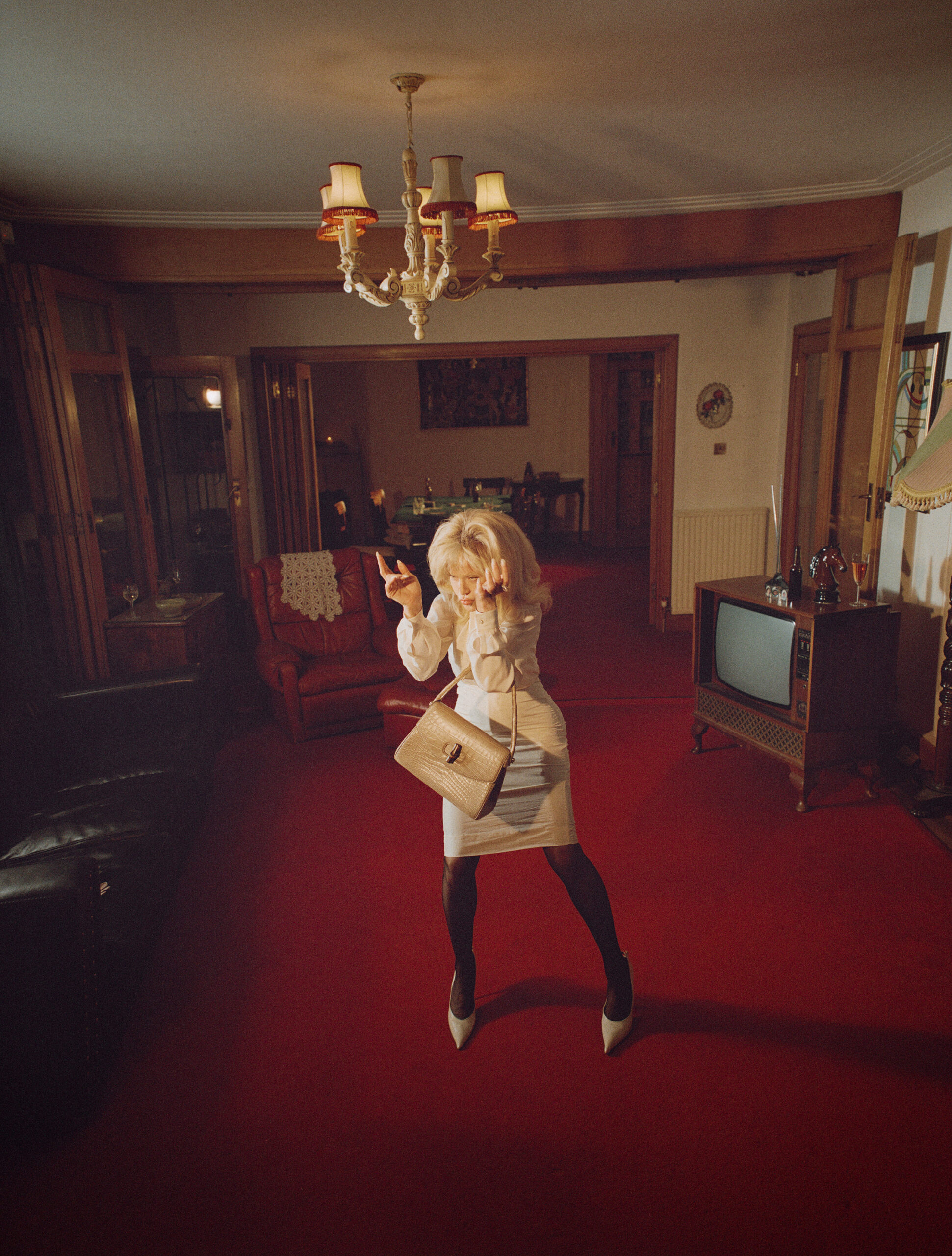 Madonna posing in a cream dress and blonde wig holding a handbag in a retro living room with red carpet.