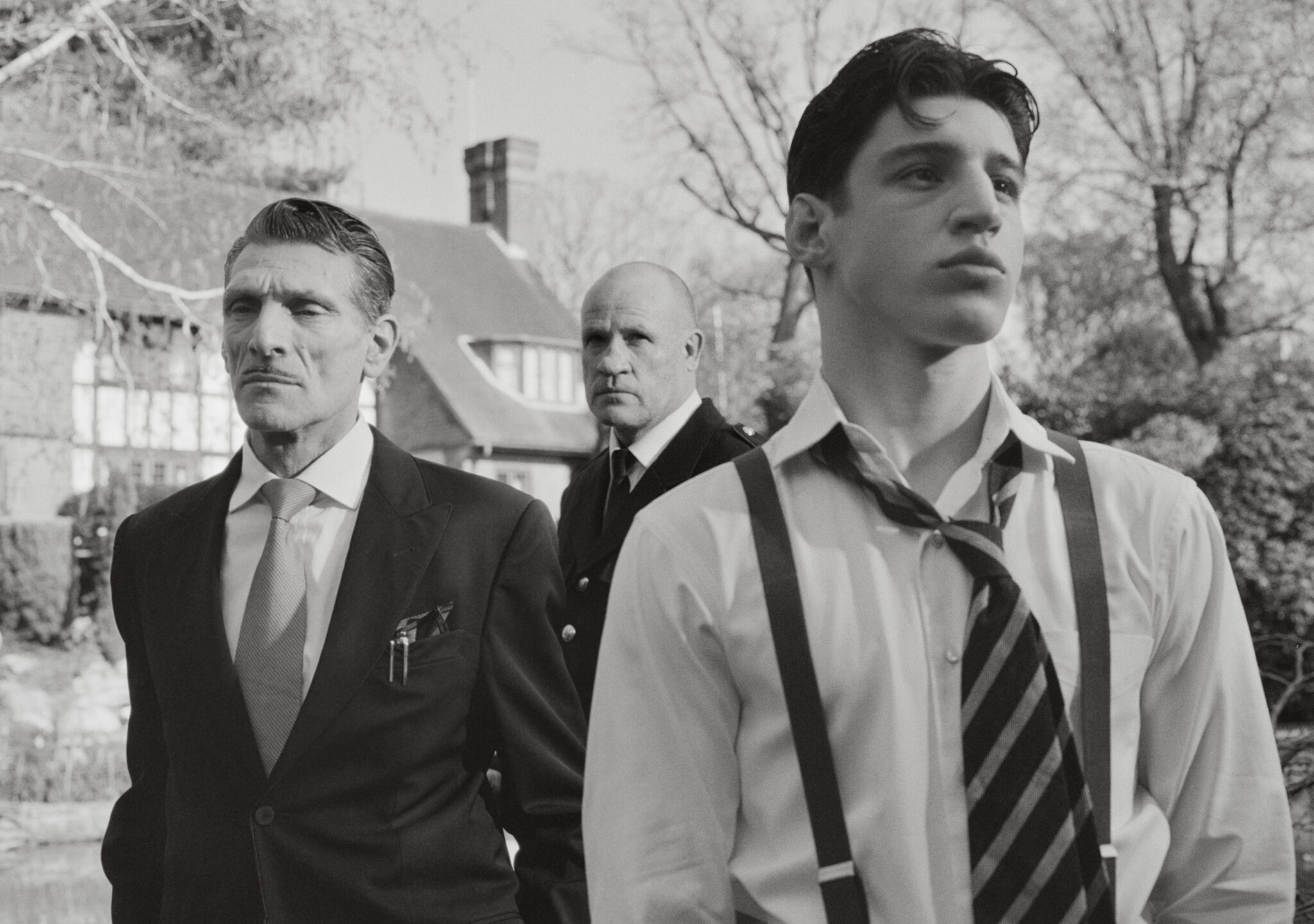 Three men in formal attire outdoors with a house in the background, photographed in black and white by Nadia Lee Cohen for Interview Magazine.