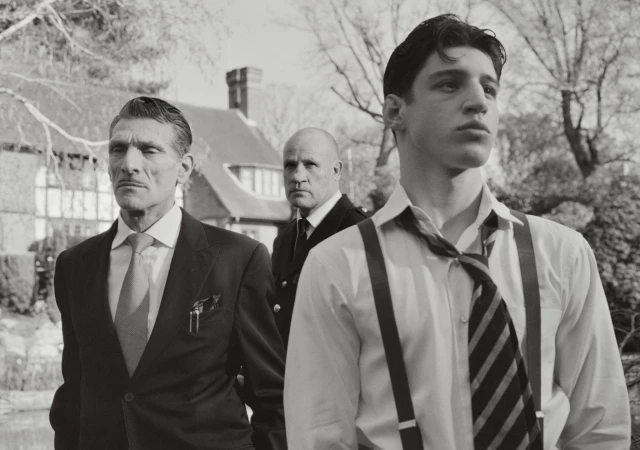 Three men in formal attire outdoors with a house in the background, photographed in black and white by Nadia Lee Cohen for Interview Magazine.