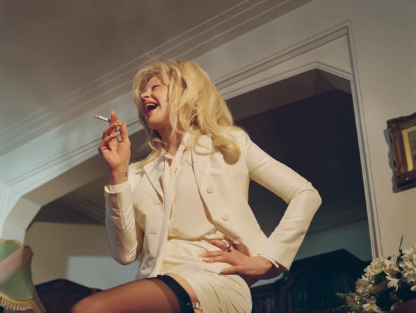 Madonna laughing with a cigarette in hand, wearing a cream blazer and skirt suit in a retro-styled domestic interior.