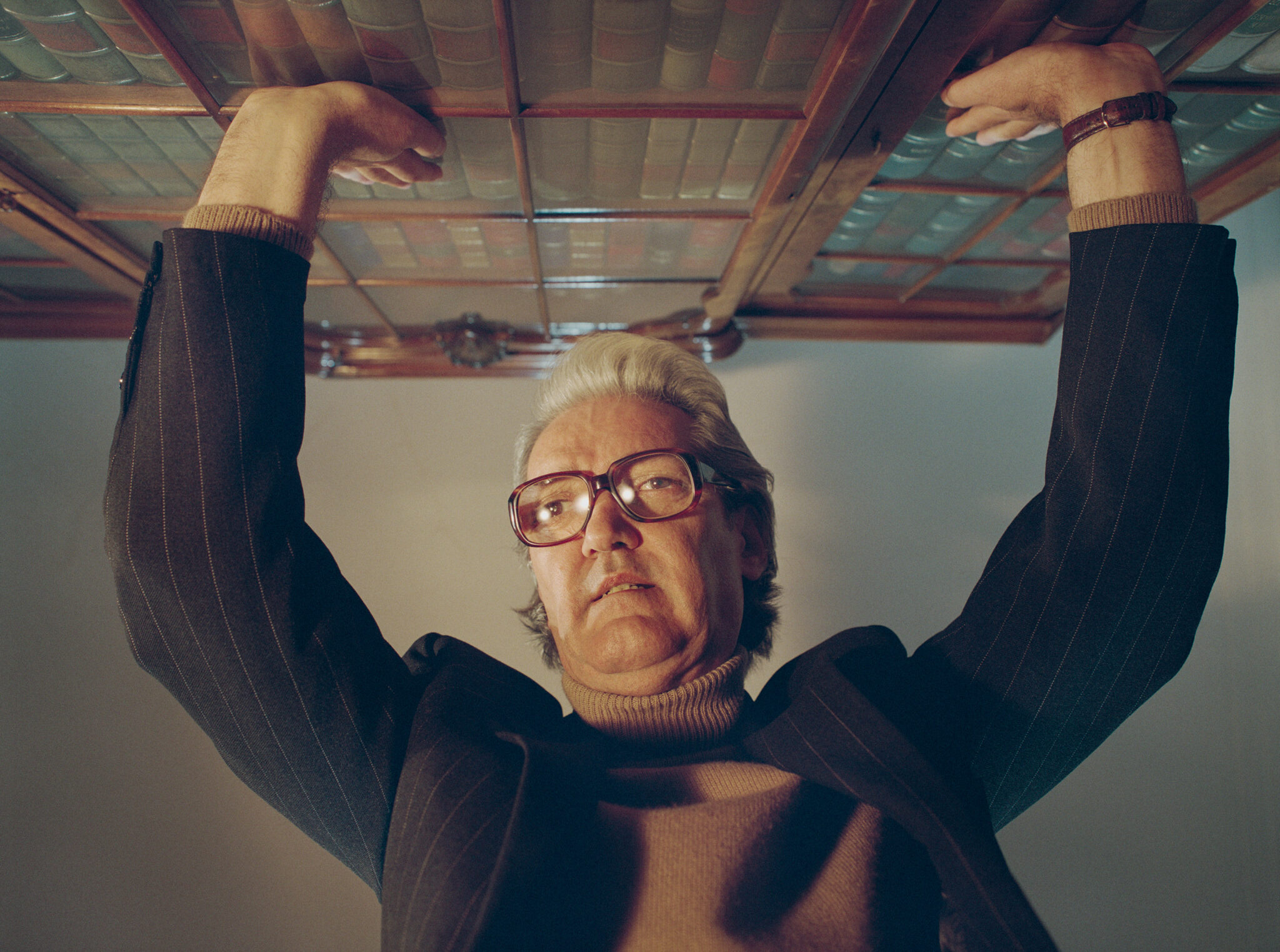 Low-angle shot of a man in glasses, pinstripe jacket, and turtleneck holding up a wooden glass-paneled bookshelf overhead.