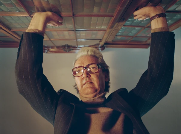 Low-angle shot of a man in glasses, pinstripe jacket, and turtleneck holding up a wooden glass-paneled bookshelf overhead.