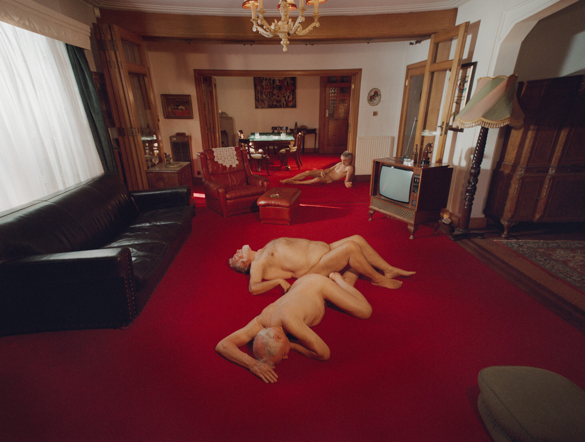 Two nude men lying on a vibrant red carpet in a retro living room with a dark leather couch and vintage television set.