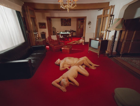 Two nude men lying on a vibrant red carpet in a retro living room with a dark leather couch and vintage television set.
