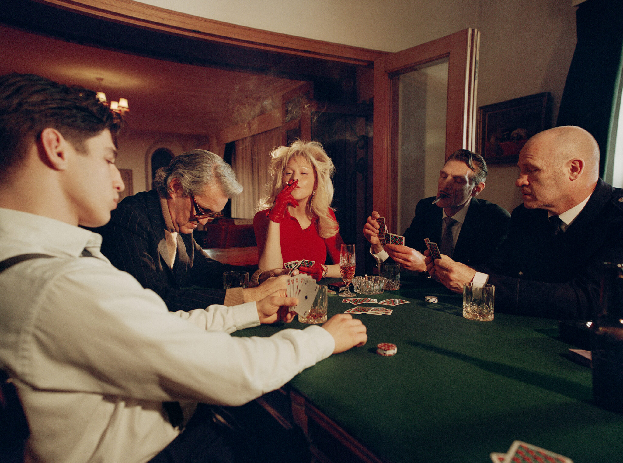 Group of people playing cards around a green felt table with glasses of whiskey and playing cards