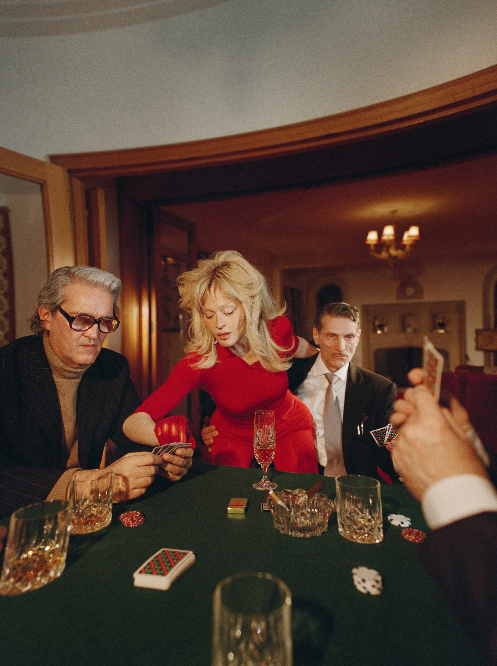 Madonna in a red dress leaning over a green poker table surrounded by men playing cards with crystal glasses and chips.