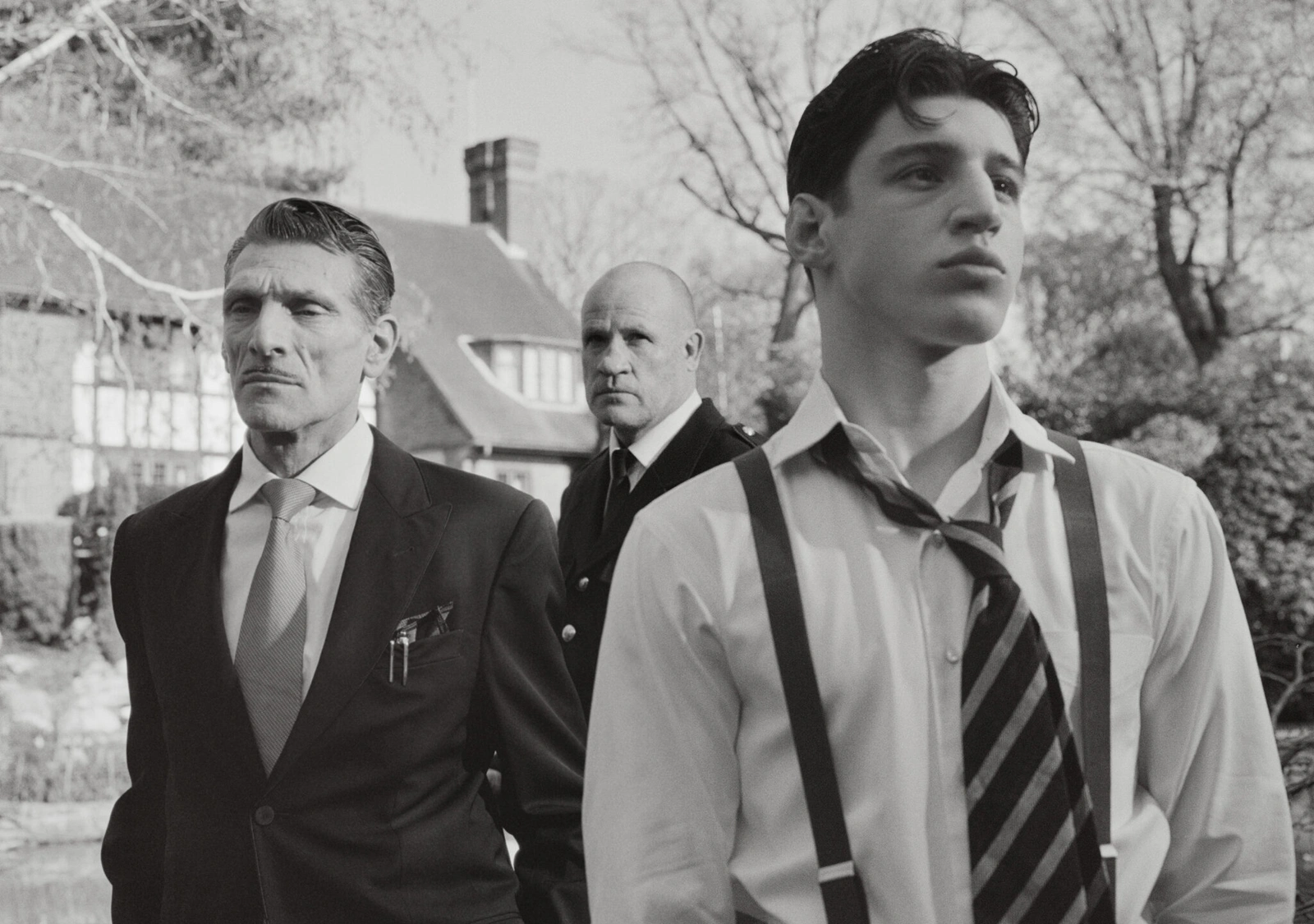 Three men in formal attire outdoors with a house in the background, photographed in black and white by Nadia Lee Cohen for Interview Magazine.