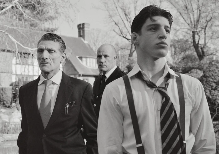 Three men in formal attire outdoors with a house in the background, photographed in black and white by Nadia Lee Cohen for Interview Magazine.