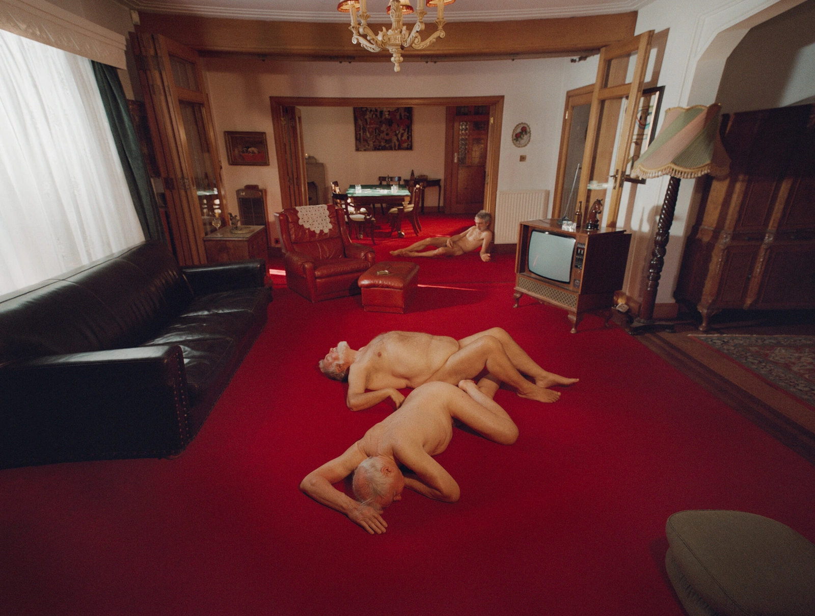 Two nude men lying on a vibrant red carpet in a retro living room with a dark leather couch and vintage television set.