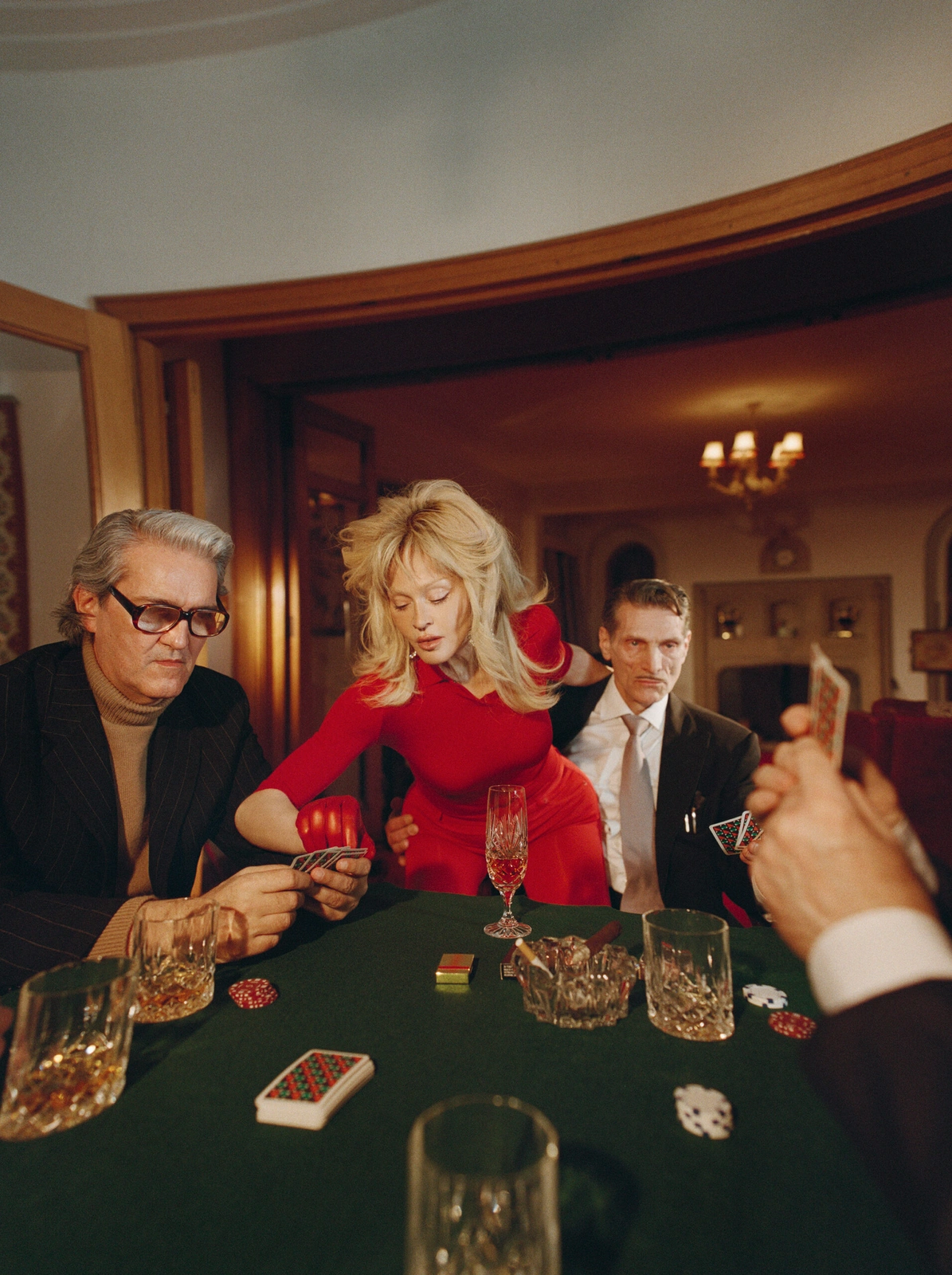 Madonna in a red dress leaning over a green poker table surrounded by men playing cards with crystal glasses and chips.