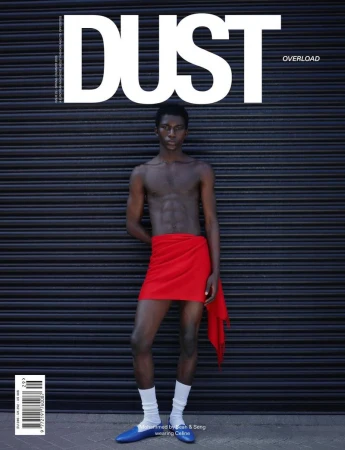 Shirtless man wearing a red wrap skirt, white socks, and blue shoes standing before a dark corrugated metal roller shutter.