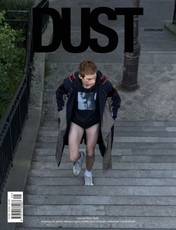 Model Joe Bottomer walks up outdoor stone stairs wearing a graphic hoodie, long coat, and sneakers for Dust Magazine.