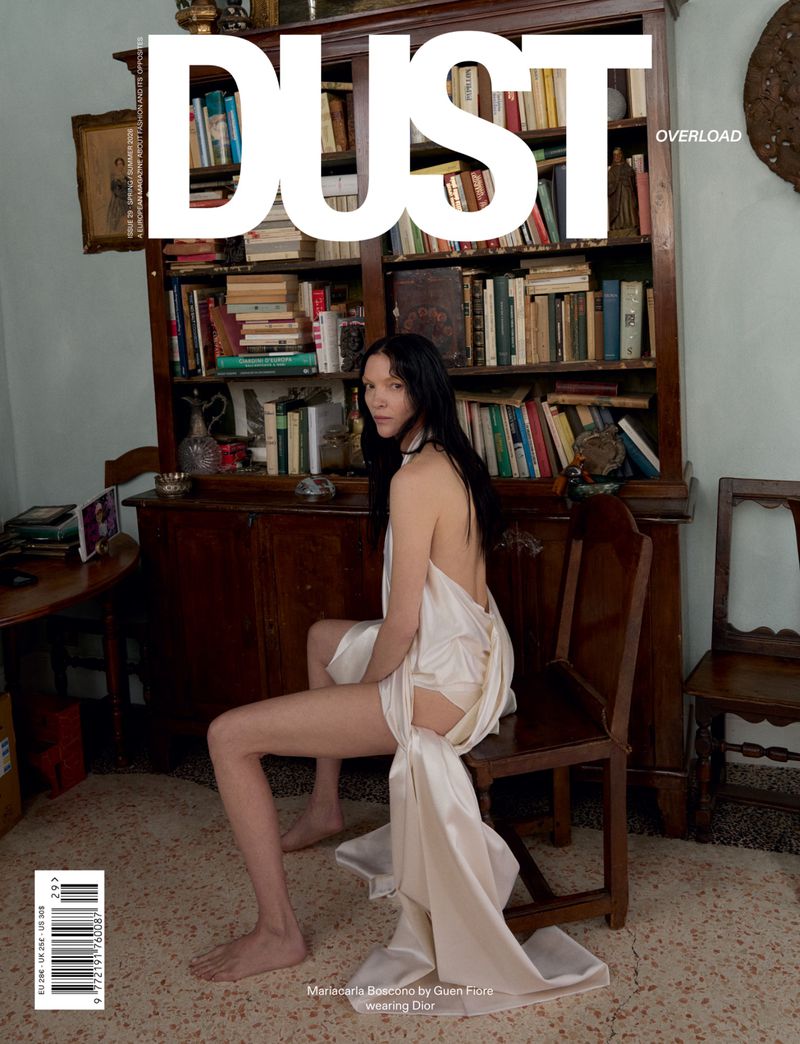 Mariacarla Boscono wearing a draped white silk backless gown seated on a wooden chair in front of a full bookshelf.