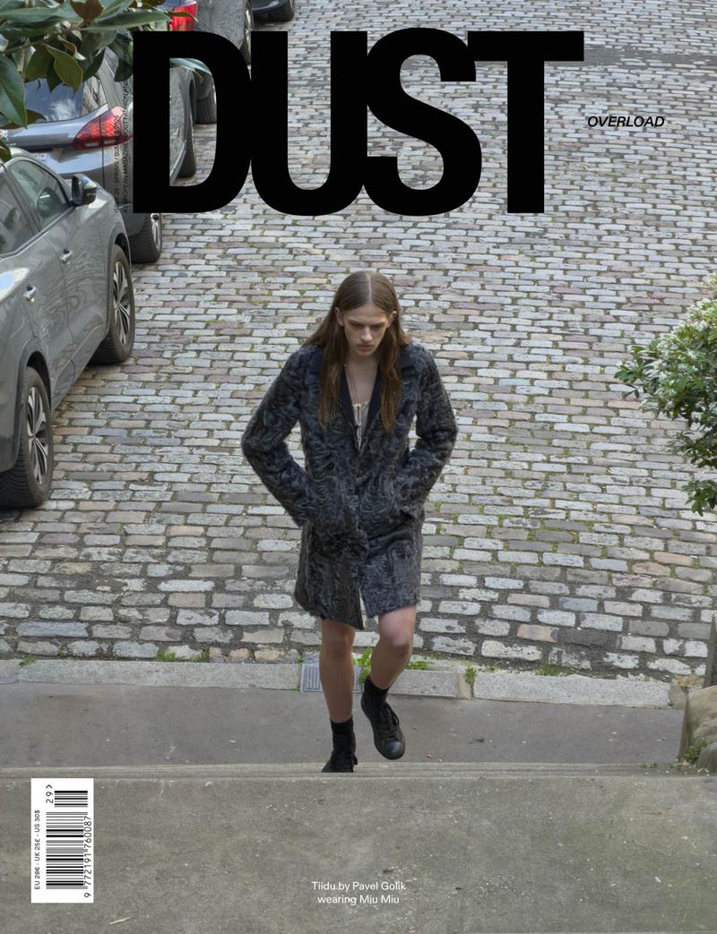 Model wearing a dark textured patterned coat walking up a cobblestone street on the cover of Dust Magazine.