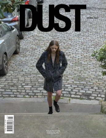 Model wearing a dark textured patterned coat walking up a cobblestone street on the cover of Dust Magazine.