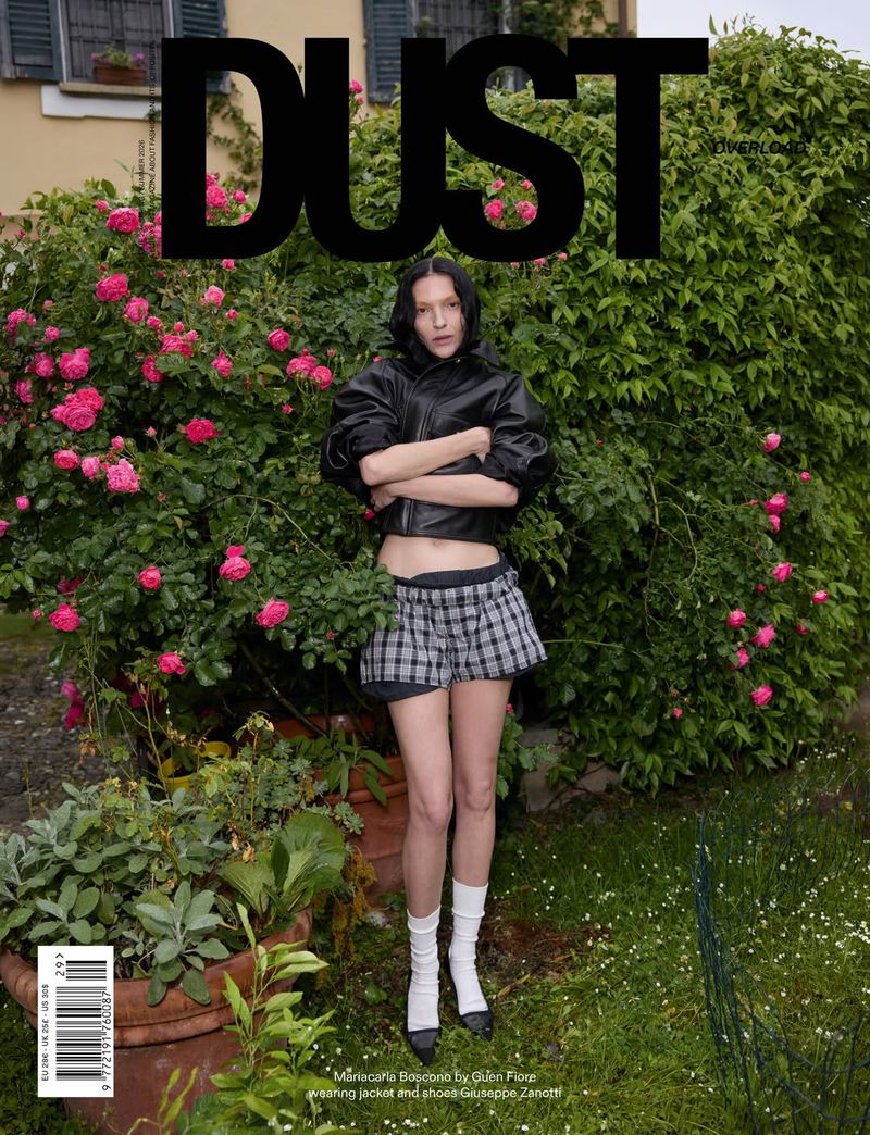 Model standing with arms crossed in front of a blooming rose bush wearing a black leather jacket and plaid skirt