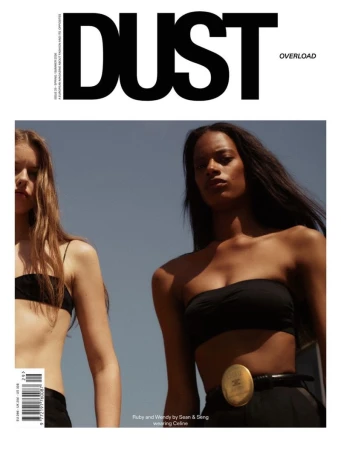 Two models outdoors under a blue sky, featuring a Black woman wearing a black bandeau top and trousers with a gold oval belt buckle.
