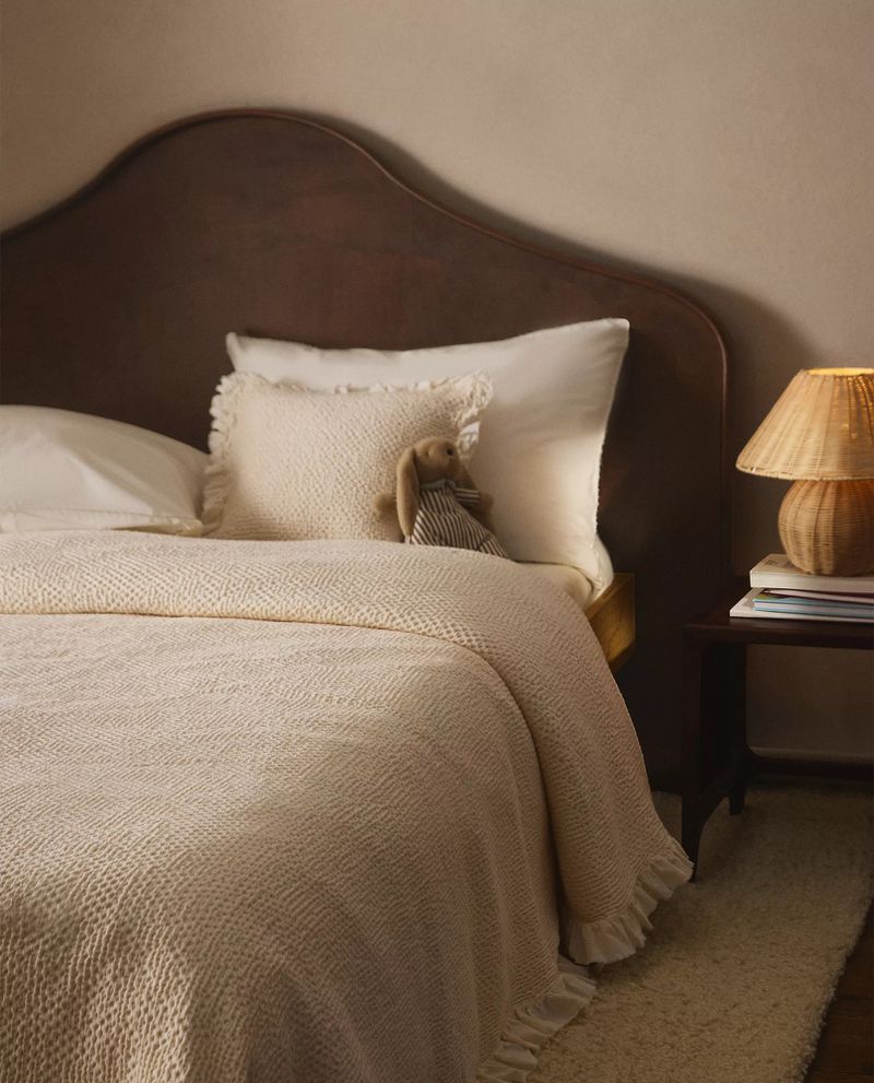 A bed with a dark curved headboard styled with cream ruffled waffle bedding, a stuffed rabbit, and a bedside lamp.
