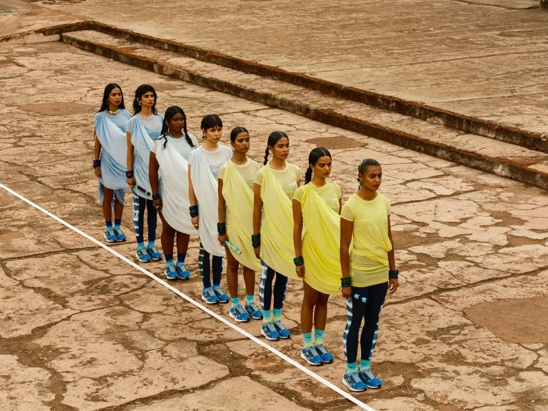 A diverse group of young women in colorful draped outfits line up outdoors on a cracked surface, showcasing contemporary fashion and style.