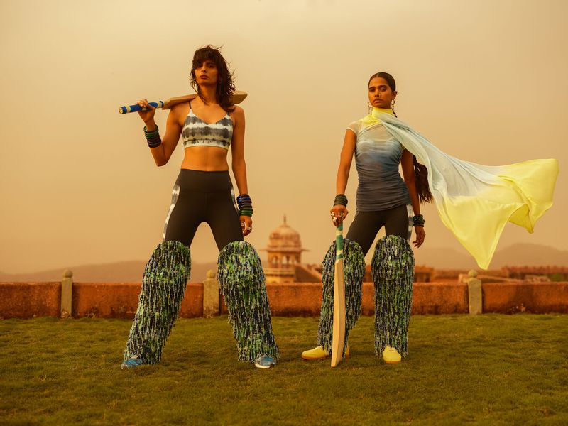 Two women in athletic wear stand with cricket bats on a rooftop, set against a hazy sunset backdrop with distant architecture.