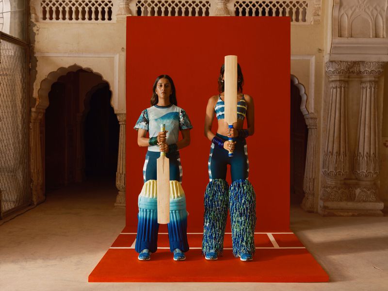 Two women in cricket attire hold bats against a red backdrop within a historic building, blending sport and cultural heritage in fashion.