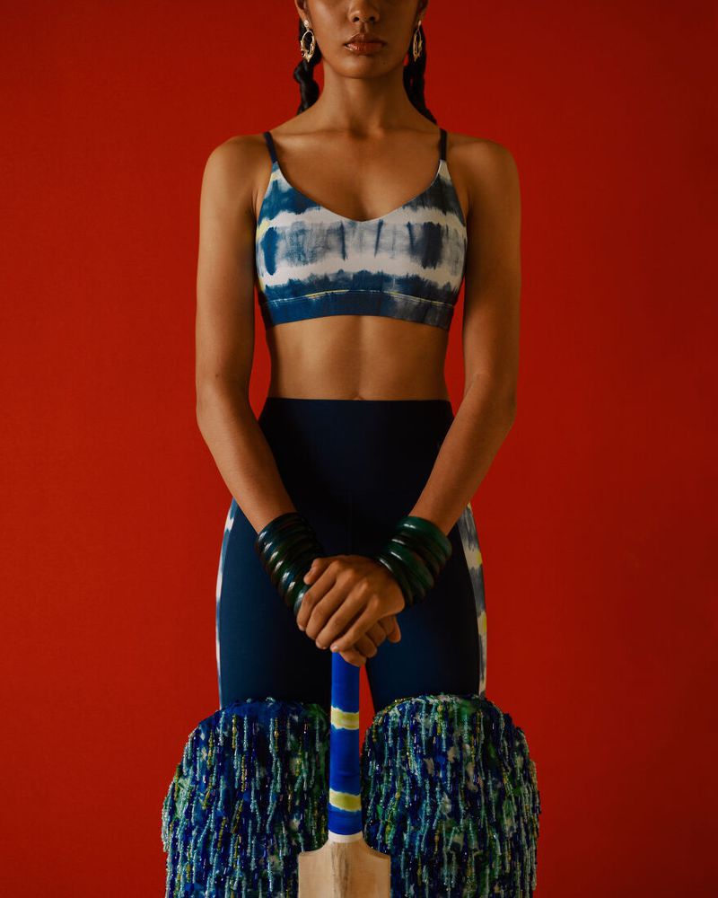 Athletic woman poses with cricket bat against a red backdrop. She wears a crop top, leggings, and bracelets. Sports and fashion fusion.