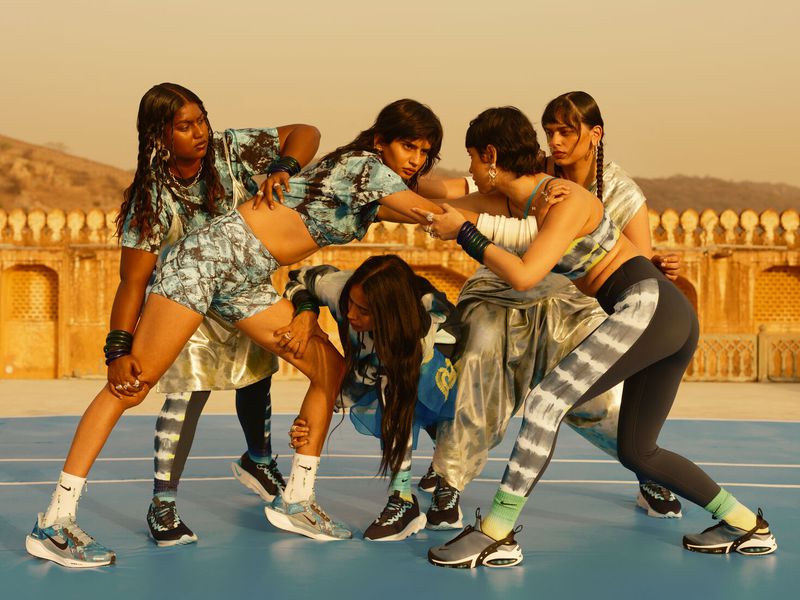 Group of five women in athletic wear pose dynamically on a blue surface with blurred landscape featuring a gold-toned architecture.