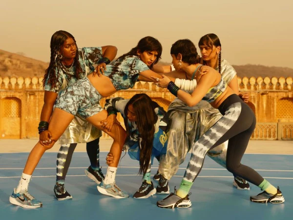 Group of five women in athletic wear pose dynamically on a blue surface with blurred landscape featuring a gold-toned architecture.