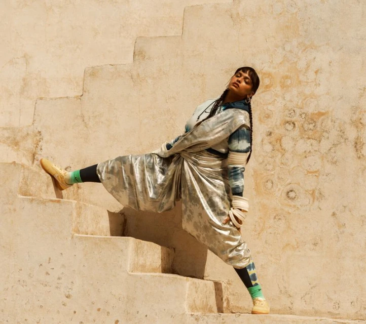 Stylish woman in metallic outfit poses on sunlit steps, showcasing fashion and urban style against a textured beige wall backdrop.