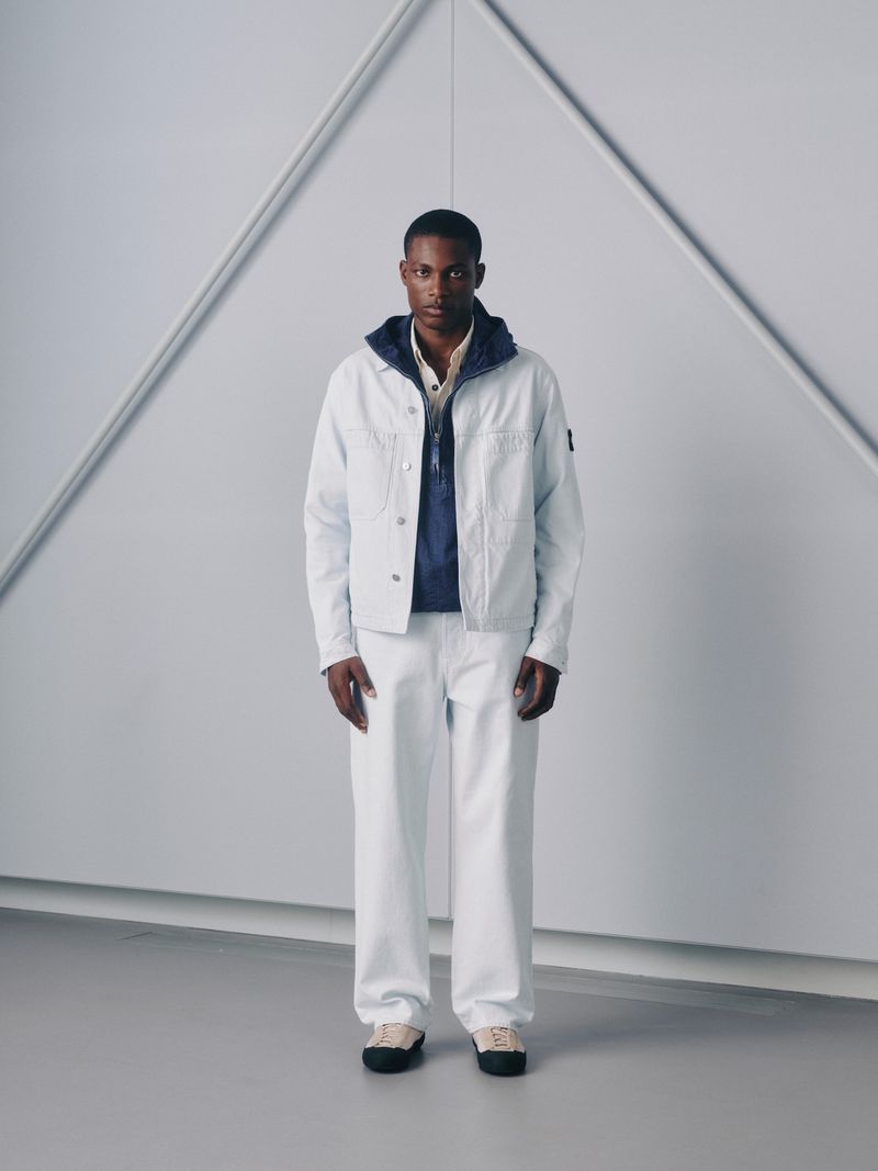 Male fashion model wears an off-white jacket and pants, in a full-length studio shot with a gray geometric backdrop and neutral sneakers.